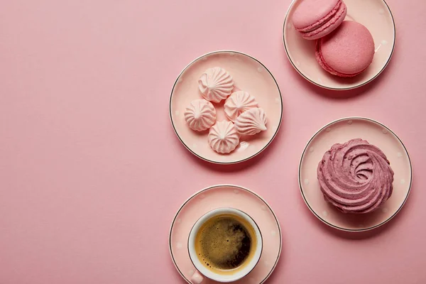 Top view of pink meringues and macaroons on small pink saucers with white dots and cup of coffee on pink background — Stock Photo