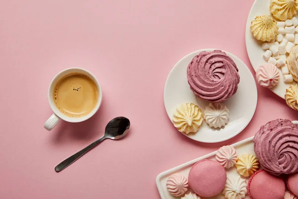 Top view of pink, white and yellow meringues, macaroons, marshmallows and cup of coffee with spoon on pink background — Stock Photo