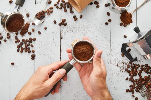 Vista superior de las manos masculinas con portafilter bajo superficie de madera blanca con cucharas, taza de papel y cafetera géiser - foto de stock