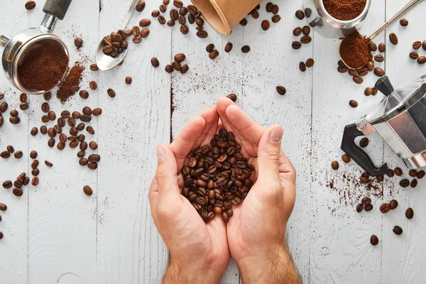 Top view of male hands with coffee beans under white wooden surface with spoons, paper cup, portafilter and geyser coffee maker — Stock Photo