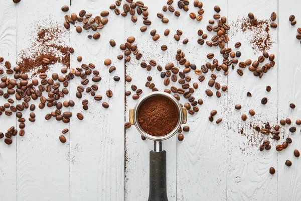 Vue du dessus du portafilter avec du café moulu sur une surface en bois blanc avec des grains de café éparpillés — Photo de stock