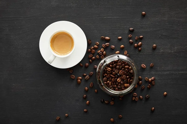 Top view of white cup of coffee on saucer near glass jar with coffee beans — Stock Photo
