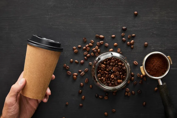 Vue de dessus de la main avec tasse en papier sous une surface en bois sombre avec bocal en verre et portafilter — Photo de stock