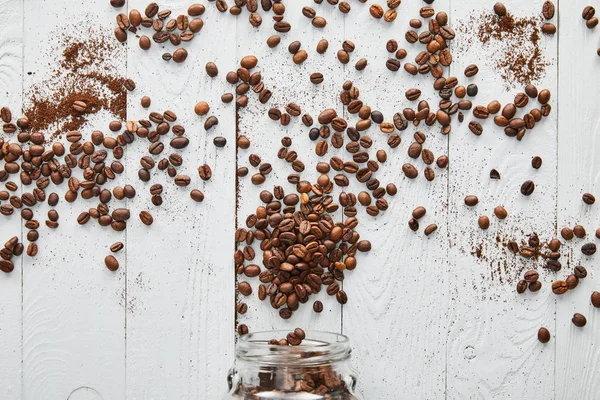 Grains de café dispersés sur la surface en bois blanc avec pot en verre — Photo de stock