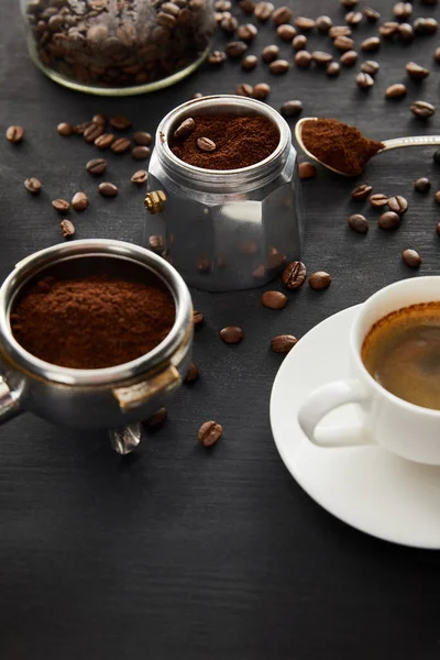 Partie de la cafetière geyser près de la tasse de café sur la soucoupe, portafilter et cuillère sur la surface en bois sombre avec des grains de café — Photo de stock