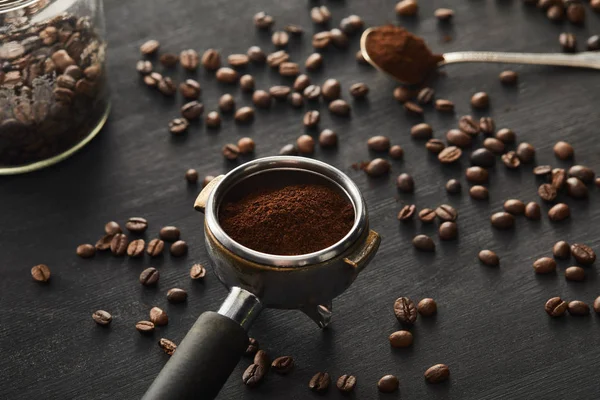 Portafilter rempli de café moulu près cuillère ang pot en verre sur une surface en bois sombre avec des grains de café — Photo de stock