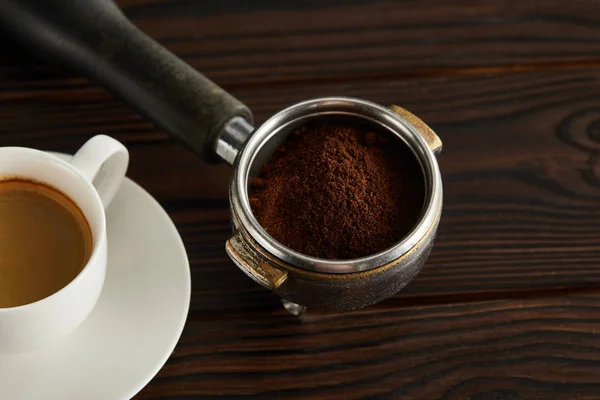 Portafilter with fresh ground coffee near cup of espresso on dark wooden surface — Stock Photo