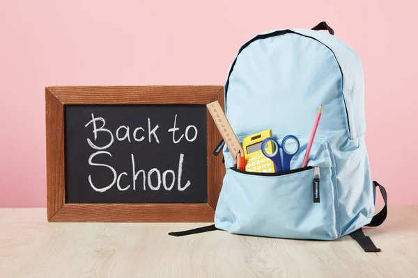 Mochila azul escolar con suministros en el bolsillo cerca de pizarra con letras de vuelta a la escuela aisladas en rosa - foto de stock