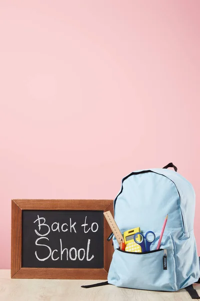 Mochila azul com suprimentos no bolso perto de quadro-negro com letras de volta para a escola isolado em rosa — Fotografia de Stock