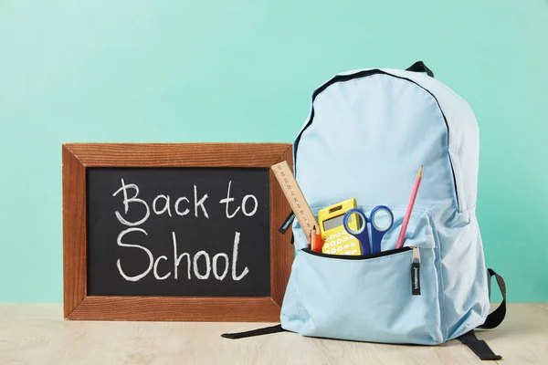 Mochila azul con suministros en el bolsillo cerca de pizarra con letras de vuelta a la escuela aisladas en turquesa - foto de stock