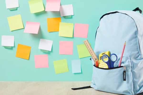 Blue backpack with supplies in pocket near multicolored sticky notes on turquoise wall — Stock Photo