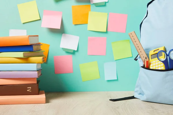 Mochila azul con suministros en el bolsillo cerca de libros y notas adhesivas multicolores en la pared turquesa - foto de stock