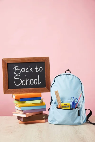 Mochila azul con suministros en el bolsillo cerca de pizarra con letras de vuelta a la escuela en libros aislados en rosa - foto de stock