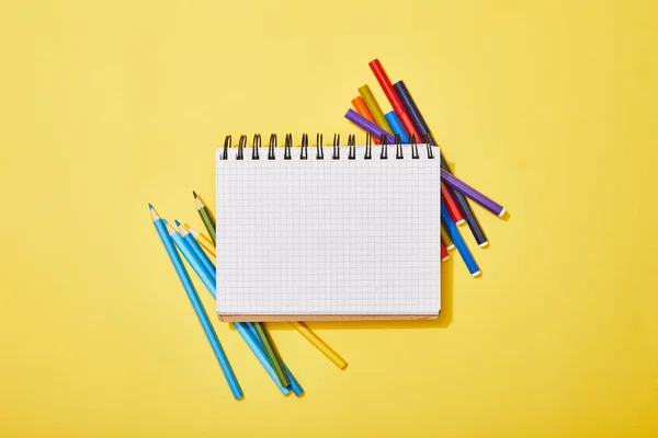 Vue du haut des crayons de couleur et des stylos en feutre éparpillés près du bloc-notes vierge sur jaune — Photo de stock