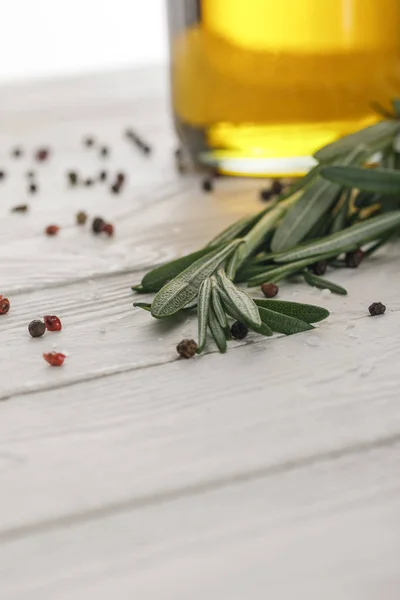 Rosemary bundle near bottle with oil and scattered red and black peppercorns on white wooden surface — Stock Photo