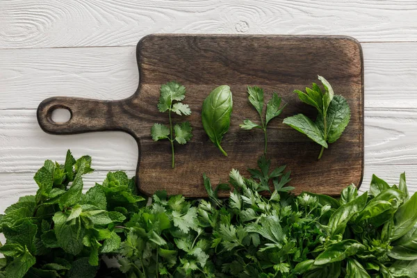 Vista superior da placa de corte de madeira marrom com manjericão, salsa, coentro e folhas de hortelã-pimenta perto de feixes de vegetação na superfície branca — Fotografia de Stock