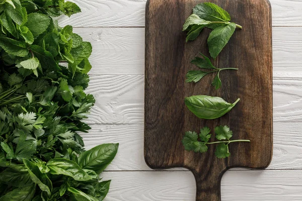 Top view of brown wooden cutting board with parsley, basil, cilantro and peppermint leaves near bundles of greenery on white surface — Stock Photo