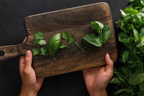 Vue recadrée de l'homme tenant une planche à découper en bois brun avec du basilic, du persil, de la coriandre et des feuilles de menthe poivrée près de faisceaux de verdure sur une surface blanche — Photo de stock