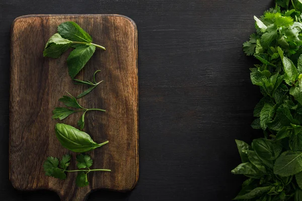 Vista superior de tabla de cortar de madera marrón con albahaca, perejil, cilantro y hojas de menta cerca de paquetes de vegetación en la superficie oscura - foto de stock