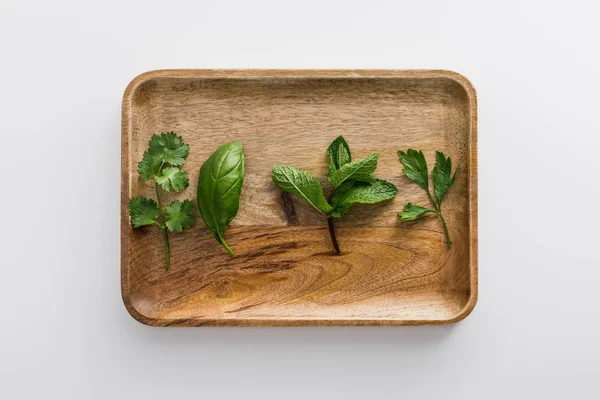 Vista dall'alto del piatto di legno marrone con prezzemolo, basilico, coriandolo e foglie di menta piperita sulla superficie bianca — Foto stock