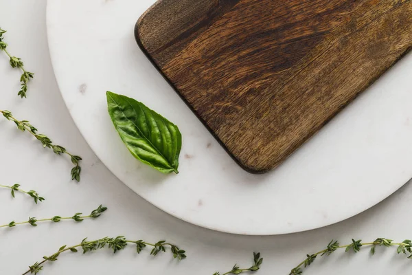 Vue de dessus de la surface ronde en marbre avec planche à découper en bois marron et feuille de basilic près des branches de thym sur fond blanc — Photo de stock