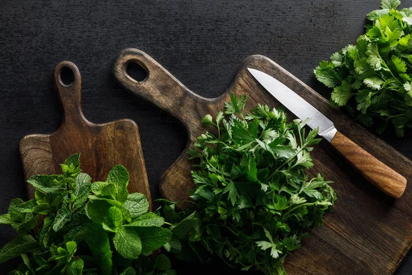 Top view of brown wooden cutting boards with parsley, peppermint and cilantro  bundles with knife on dark surface — Stock Photo