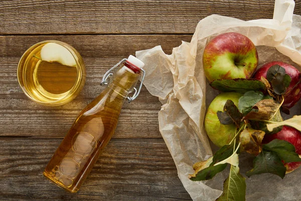Вид складной пергаментной бумаги с яблоками возле бутылки и стакана сидра на деревянной поверхности — стоковое фото