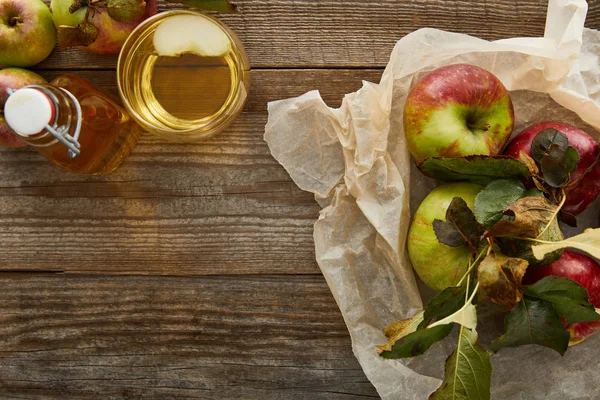 Draufsicht auf Pergamentpapier mit Äpfeln in Flaschennähe und Glas Apfelwein auf Holzoberfläche — Stockfoto