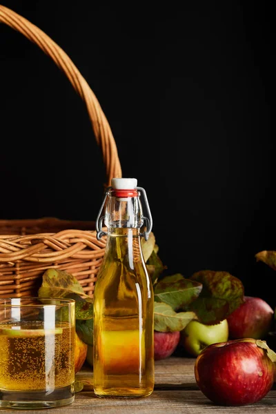 Botella y vaso de sidra cerca de la cesta de mimbre y manzanas en la superficie de madera aislado en negro - foto de stock