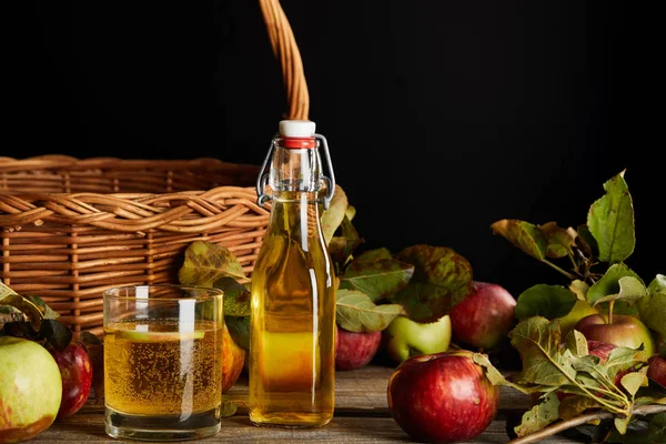 Botella y vaso de sidra casera cerca de canasta de mimbre y manzanas aisladas en negro - foto de stock