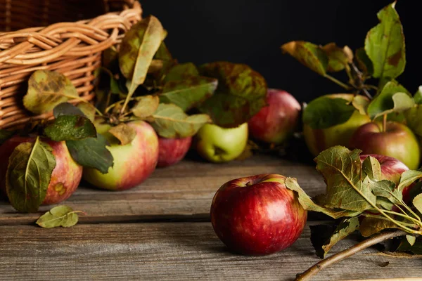 Superficie in legno con mele e foglie vicino cesto di vimini isolato su nero — Foto stock