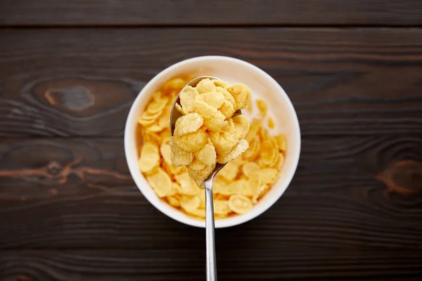 Top view of spoon with cornflakes above bowl on wooden surface — Stock Photo