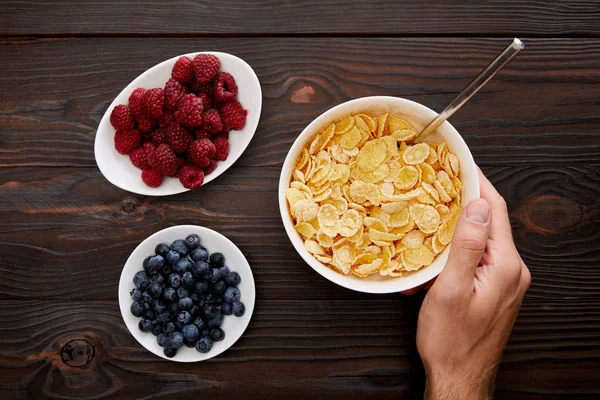 Cropped image of man holding bowl with cornflakes near plates with fresh berries on wooden surface — Stock Photo