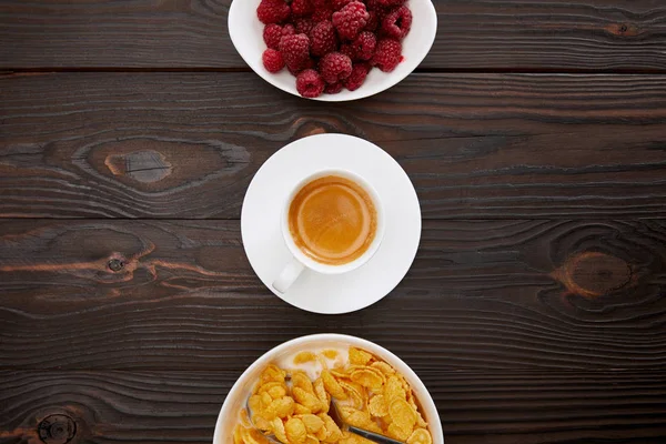 Top view of bowl with cornflakes and milk near cup of coffee and plate of raspberry on wooden surface — Stock Photo