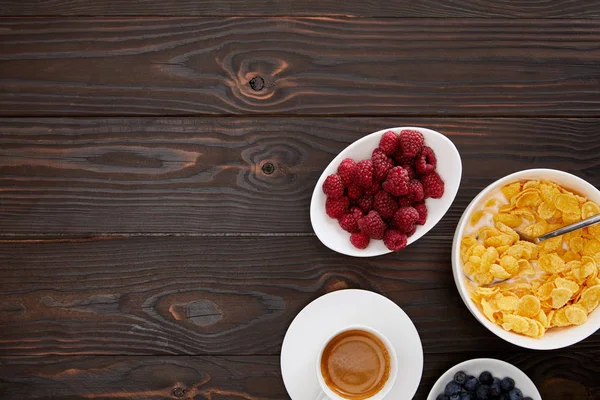 Top view of bowl with cornflakes near cup of coffee and plates of fresh berries on wooden surface — Stock Photo