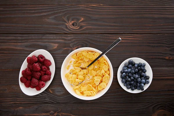 Top view of bowl with cornflakes near plates with raspberry and blueberry on wooden surface — Stock Photo