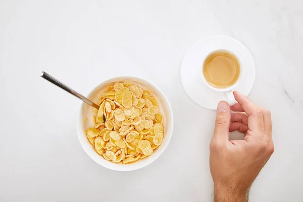 Abgeschnittenes Bild eines Mannes, der eine Tasse Kaffee in der Nähe einer Schüssel mit Cornflakes auf weißem Marmorhintergrund hält — Stockfoto