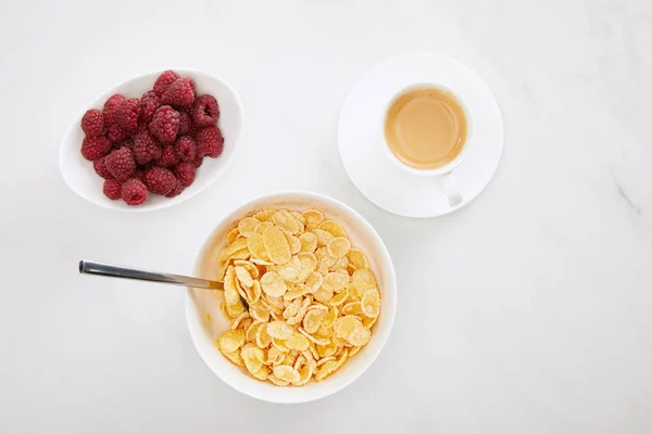 Верхний вид чаши кукурузных хлопьев с ложкой возле чашки кофе и тарелки с малиной на поверхности белого мрамора — стоковое фото