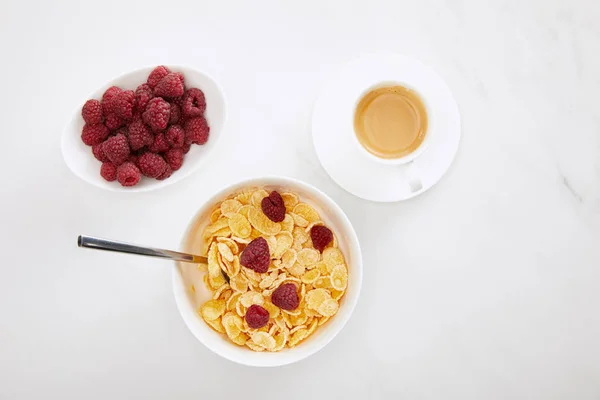 Вид зверху чаші кукурудзяних пластівців з малиною біля чашки кави та тарілки зі свіжими ягодами на поверхні білого мармуру — Stock Photo