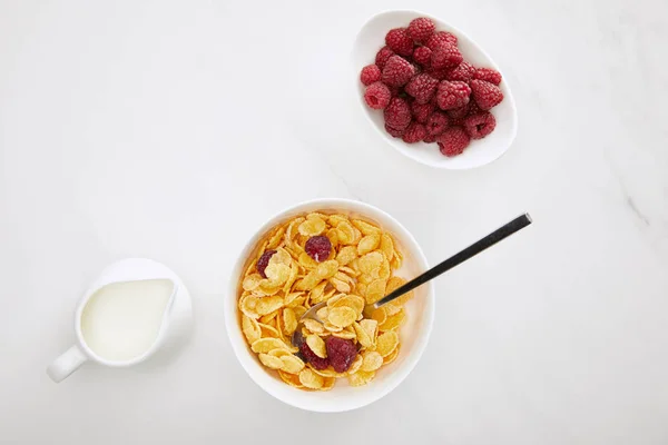 Top view of cornflakes bowl near fresh raspberry and milk jug on white marble surface — Stock Photo