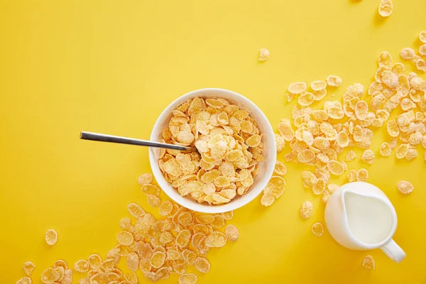 Draufsicht auf Schüssel mit Cornflakes in der Nähe von verstreuten Stücken und Krug Milch isoliert auf gelb — Stockfoto