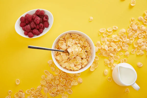 Top view of bowl with cornflakes near plate with raspberry and jug of milk isolated on yellow — Stock Photo