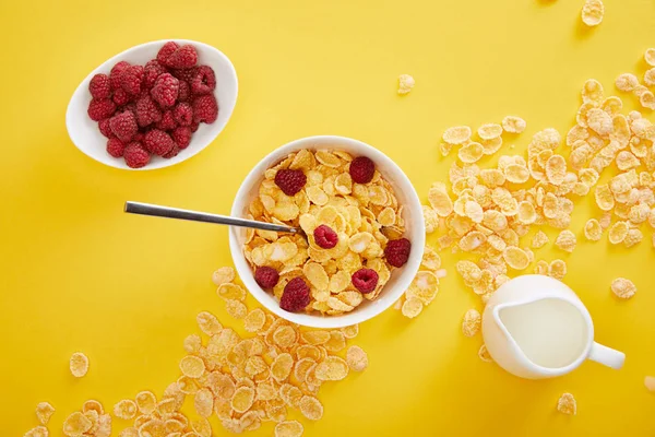 Top view of bowl with cornflakes near plate with fresh raspberry and jug of milk isolated on yellow — Stock Photo