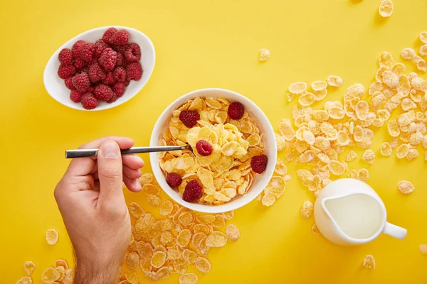 Vista recortada del hombre sosteniendo cuchara en un tazón con copos de maíz cerca de frambuesa fresca y jarra de leche aislada en amarillo - foto de stock