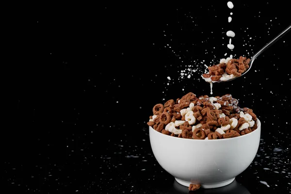 Tazón blanco con surtido de cereales y cuchara con gotas de leche aisladas en negro - foto de stock