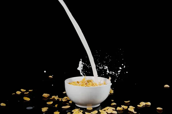 Tazón blanco con copos de maíz con chorro de leche y salpicaduras aisladas en negro - foto de stock