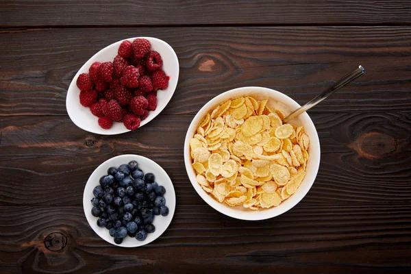 Top view of plates with raspberry and blueberry near cornflakes on wooden surface — Stock Photo