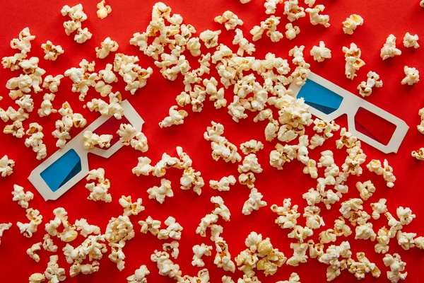 Ansicht von 3D-Gläsern auf köstlich verstreutem Popcorn auf rotem Hintergrund — Stockfoto