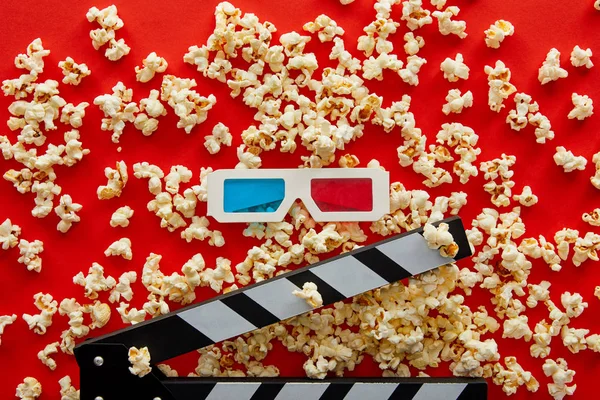 Вид сверху на вкусный попкорн, разбросанный на красном фоне возле доски хлопанья и 3D очков — стоковое фото