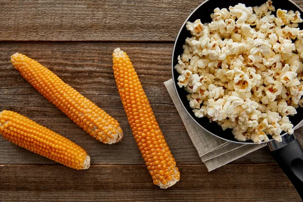 Vista dall'alto di deliziosi popcorn in padella vicino al mais su sfondo di legno — Foto stock